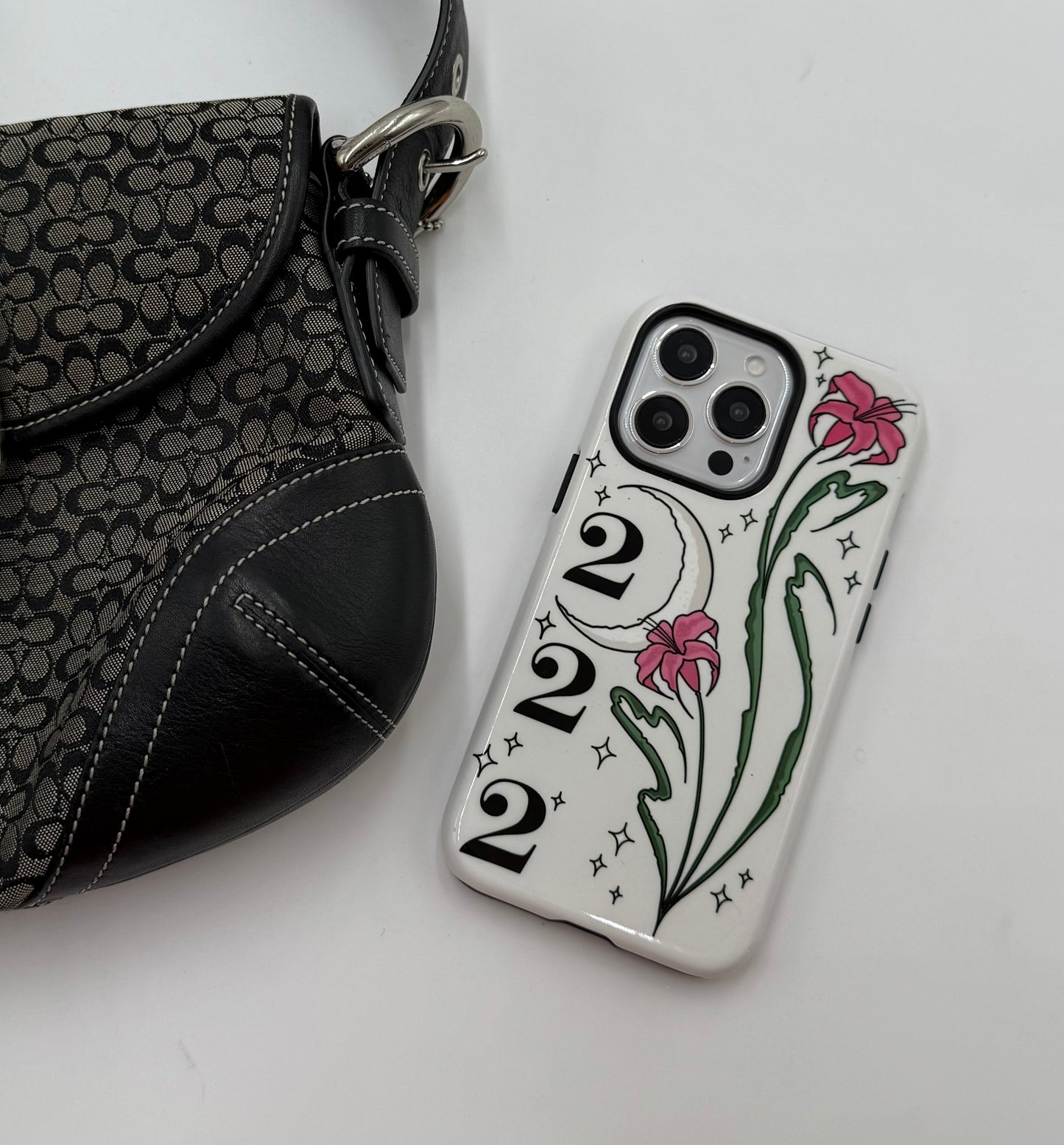 Phone case with pink lilies, crescent moon, 222 angel number on white background. twinkle stars Cute, aesthetic, protective design. Fits iPhone, Samsung & Google Pixel models. on table with bag
