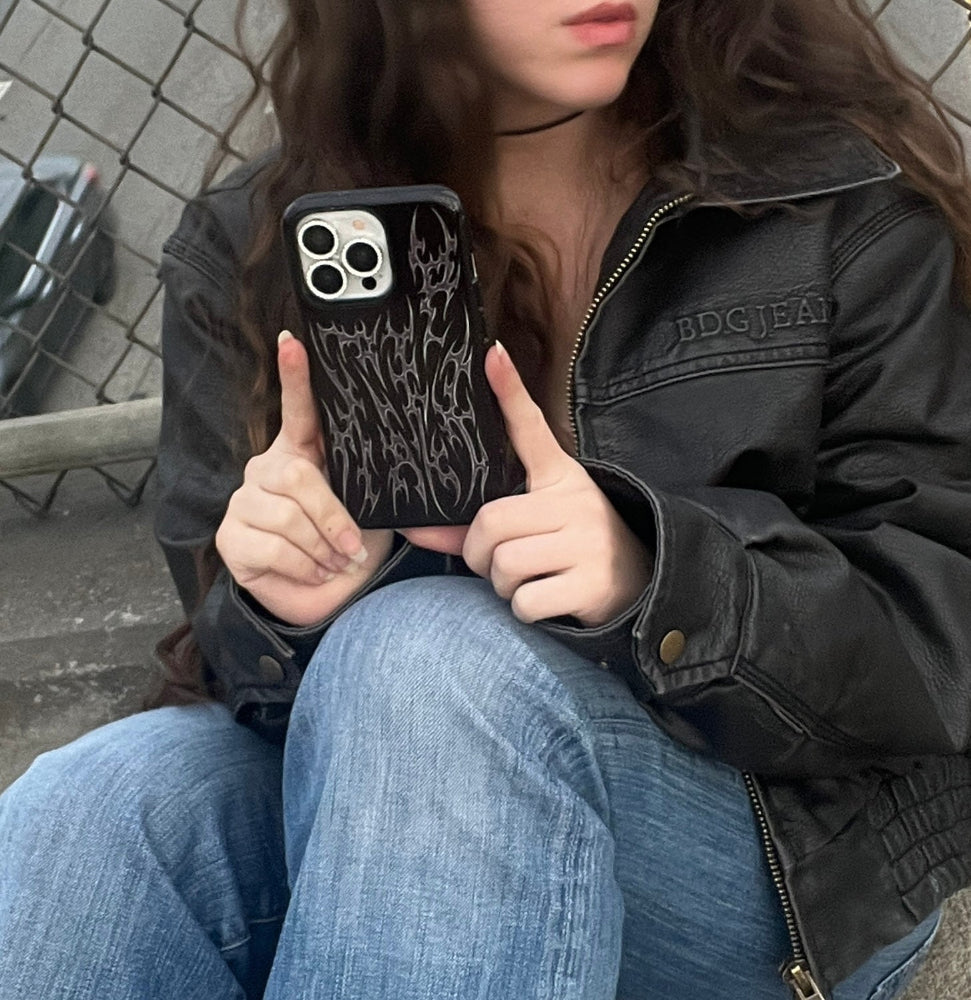 Person holding a phone with a black case featuring cyber cigilism chrome designs, wearing a black jacket and blue jeans.