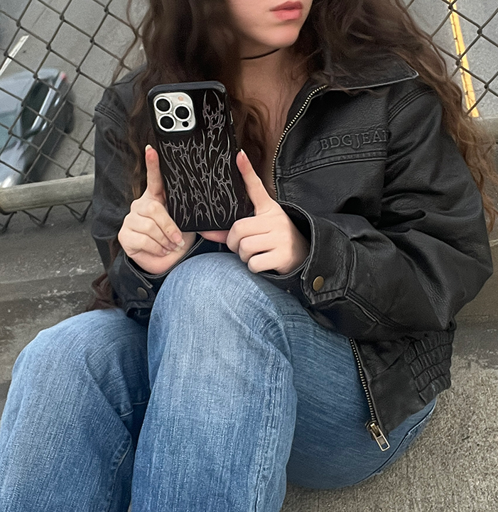 Person holding a phone with a black case, wearing a black jacket and blue jeans, sitting on steps.