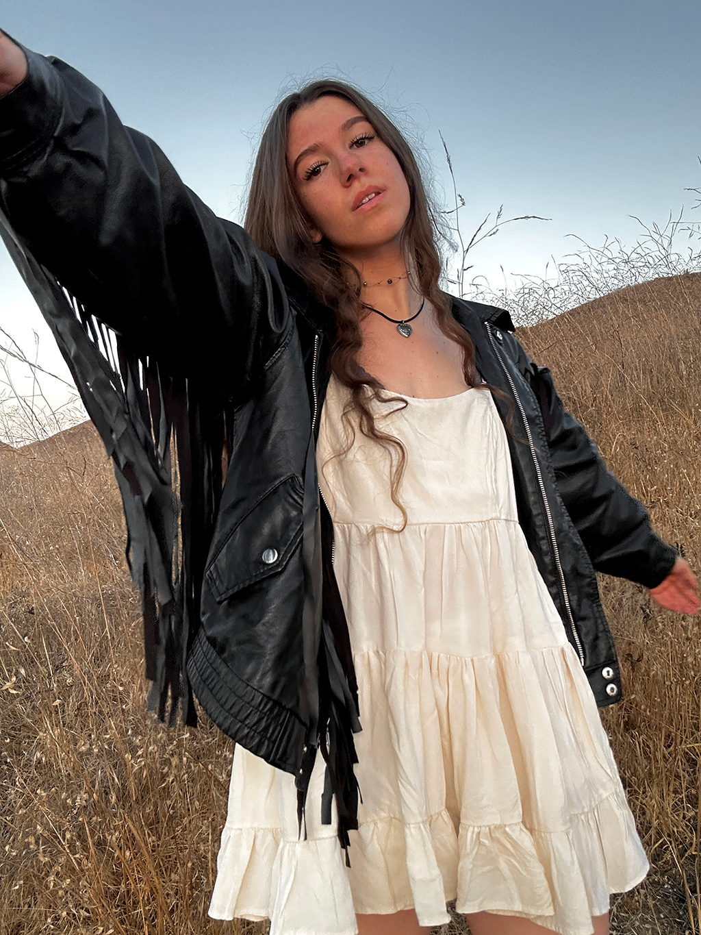 Woman in a white dress and black leather jacket standing in a field with a blue sky.