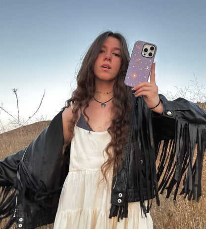 phone case with celestial sun, moon & shooting stars on purple blue background. Protective & stylish. Fits iPhone, Samsung & Google Pixel models. Boho chic, cute, and aesthetic held by model in fringe jacket