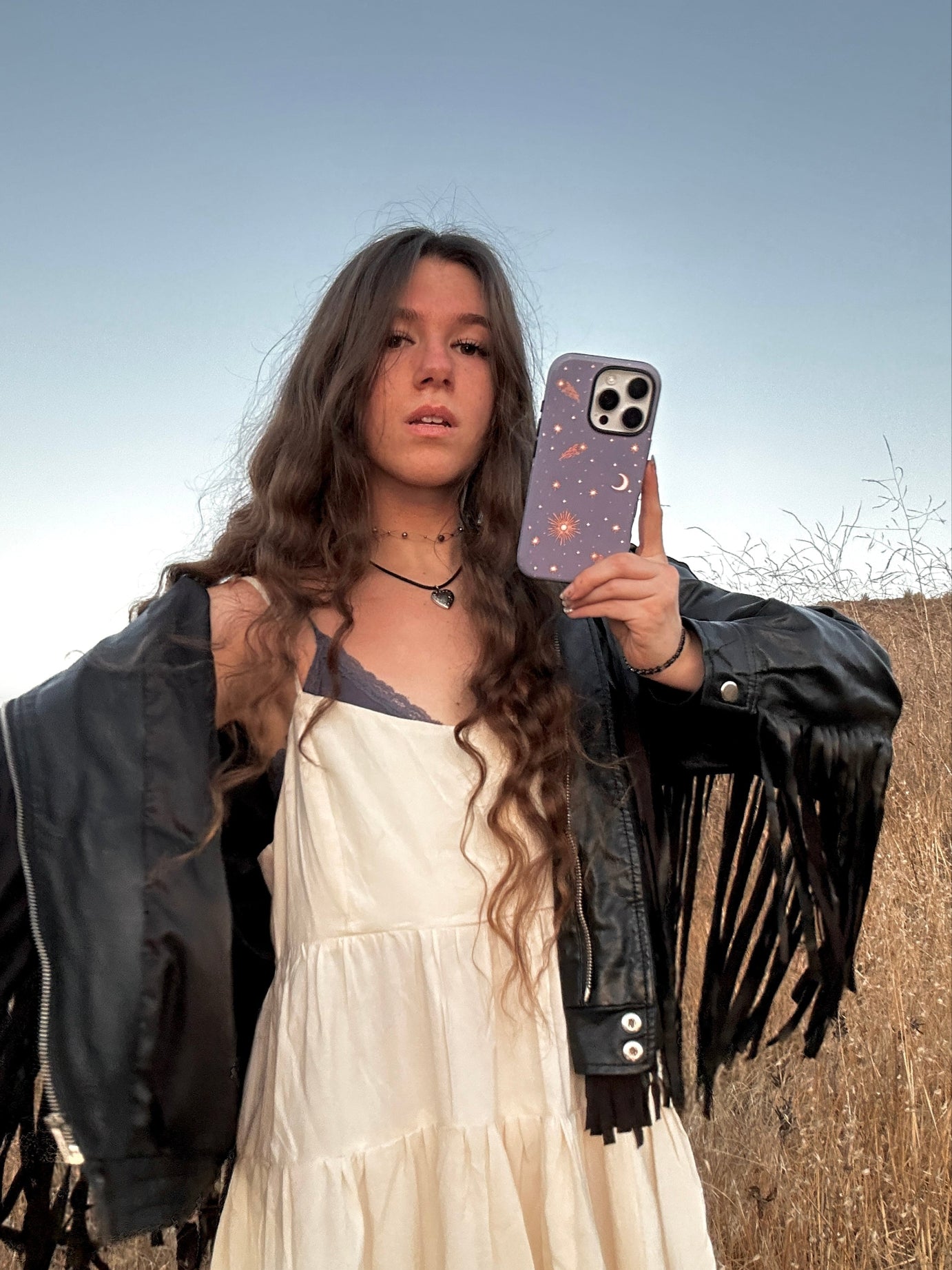 Person taking a mirror selfie outdoors wearing a white dress and black jacket with fringes.